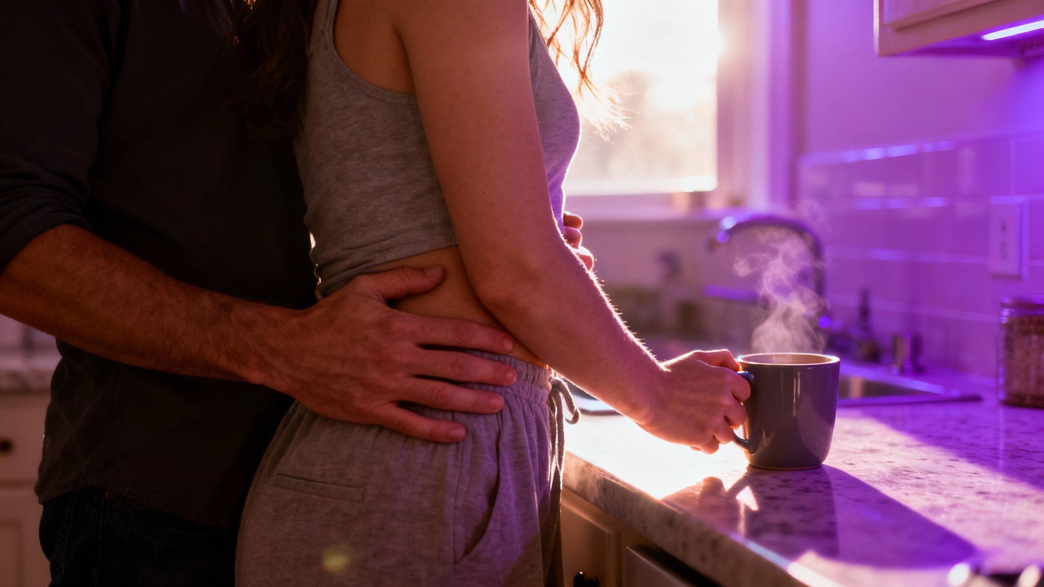 A man embraces his wife from behind in a kitchen, an example of the non-sexual 'kindling' needed to build all-day desire and please your wife in bed.